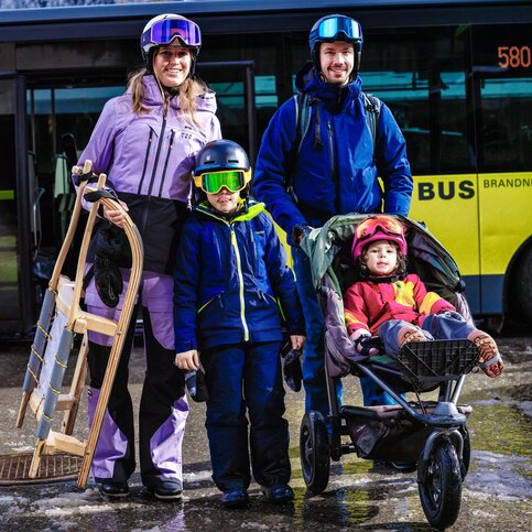 Mit dem Bus unterwegs, mit der Gästekarte kostenlos. Foto: Markus und Karin Gmeiner / Alpenregion Bludenz Tourismus Familie mit Rodel und Kinderwagen vor einem Linienbus im Winter